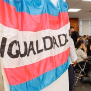 Em primeiro plano, uma bandeira transexual com o escrito "igualdade" no centro em uma porta branca. Em segundo plano, uma sala com várias pessoas sentadas em cadeiras e olhando para uma mesma direção.