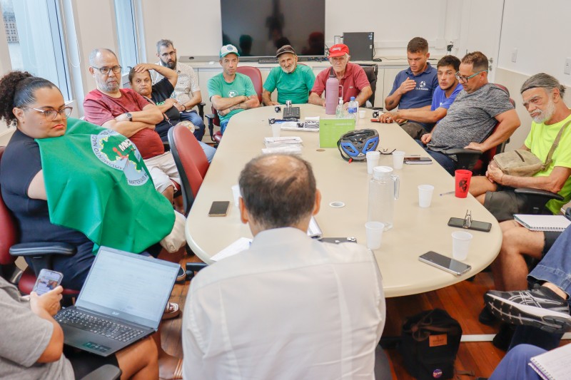 De costas para a foto, um homem está sentado a uma mesa redonda dentro de uma sala e de frente para outras 11 pessoas que, também sentadas à mesma mesa, olham para ele. 