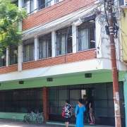 Fachado do prédio. Ele é verde com detalhes em tijolo à vista, há grades nas janelas.