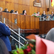 Em primeiro plano, desfocado, aparecem flores vermelhas. Ao fundo, autoridades compõe a mesa.